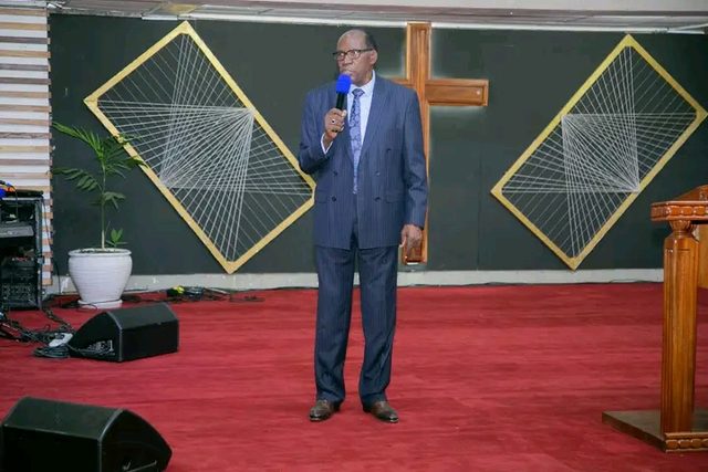 A minister in a pinstripe suit speaking beside a wooden cross
